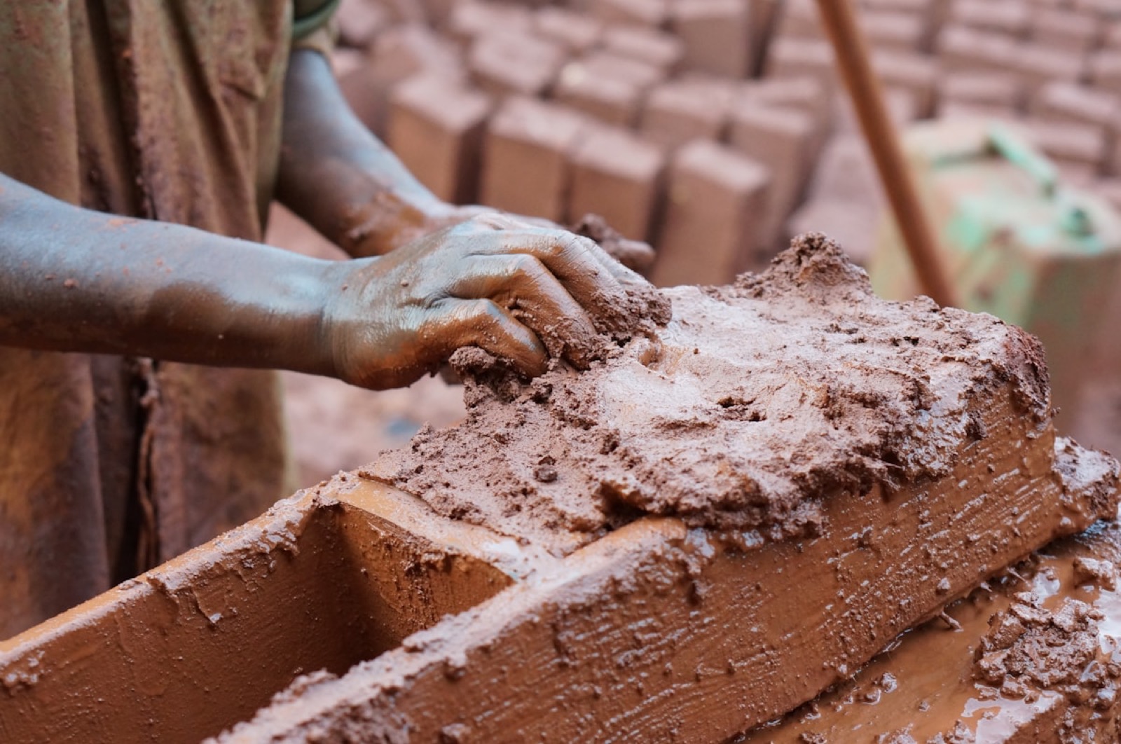 A brickmaker working clay into a wooden mould, a local brick yard stacked behind.