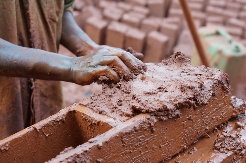 A brickmaker working clay into a wooden mould, a local brick yard stacked behind.