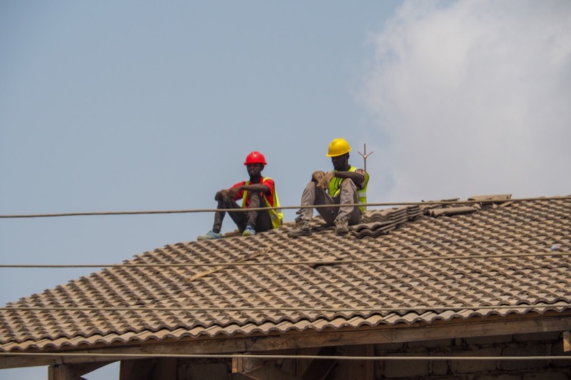 Two Ugandan workers in helmets and high-visibility vests resting on a tiled roof.